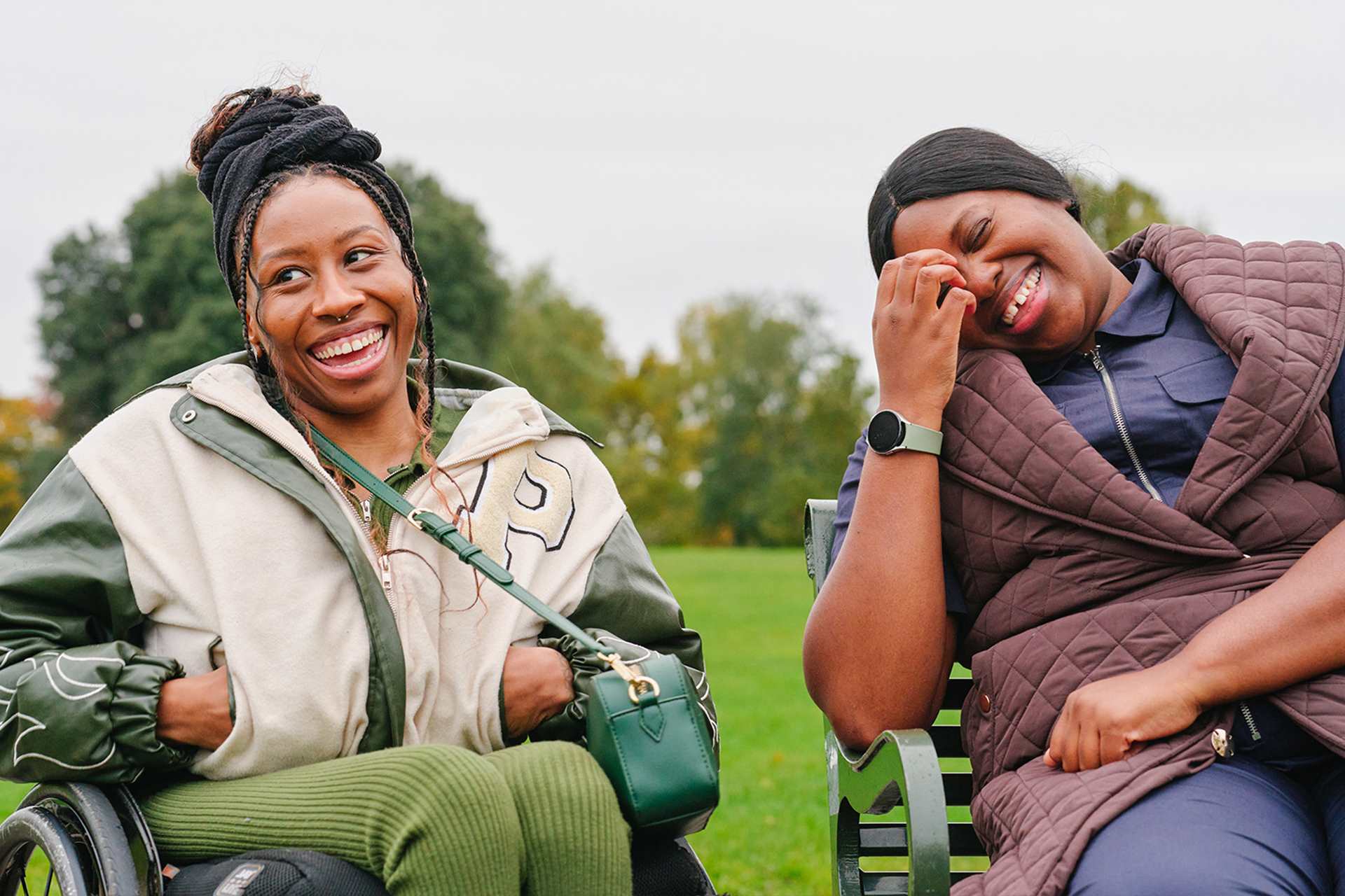 A young Black woman in a wheelchair and an older Black woman sitting on a bench in the park. They are laughing together.