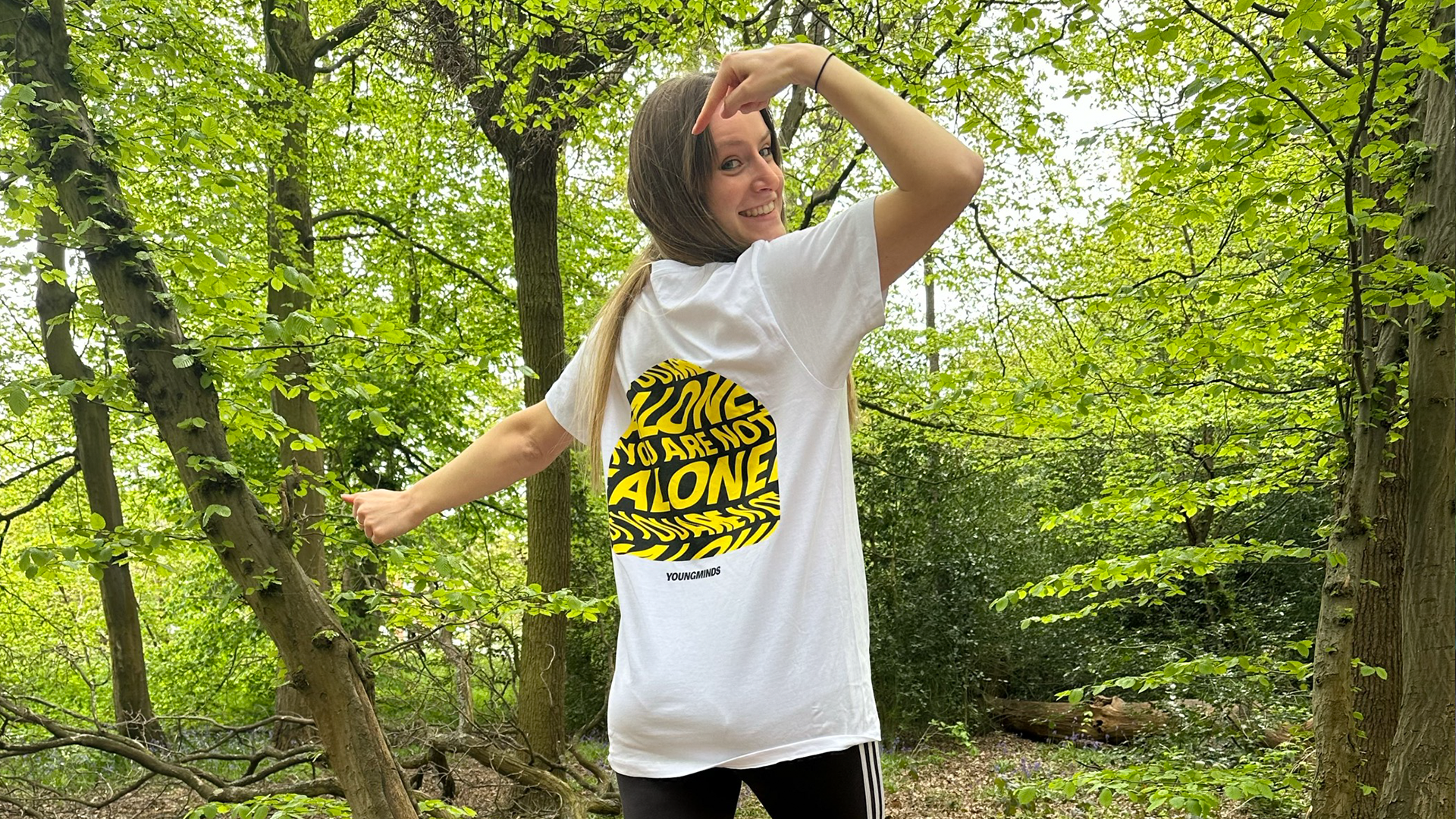A girl wearing a YoungMinds tshirt is standing in the woods. She has her back to the camera but has turned her head around and is smiling. She is pointing at the YoungMinds t-shirt logo on the back of her tshirt.