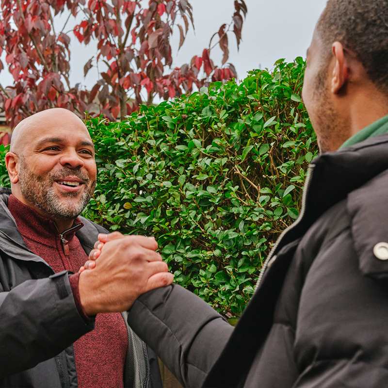 A young Black man grasping hands with an older Black man on the street. They are both smiling.