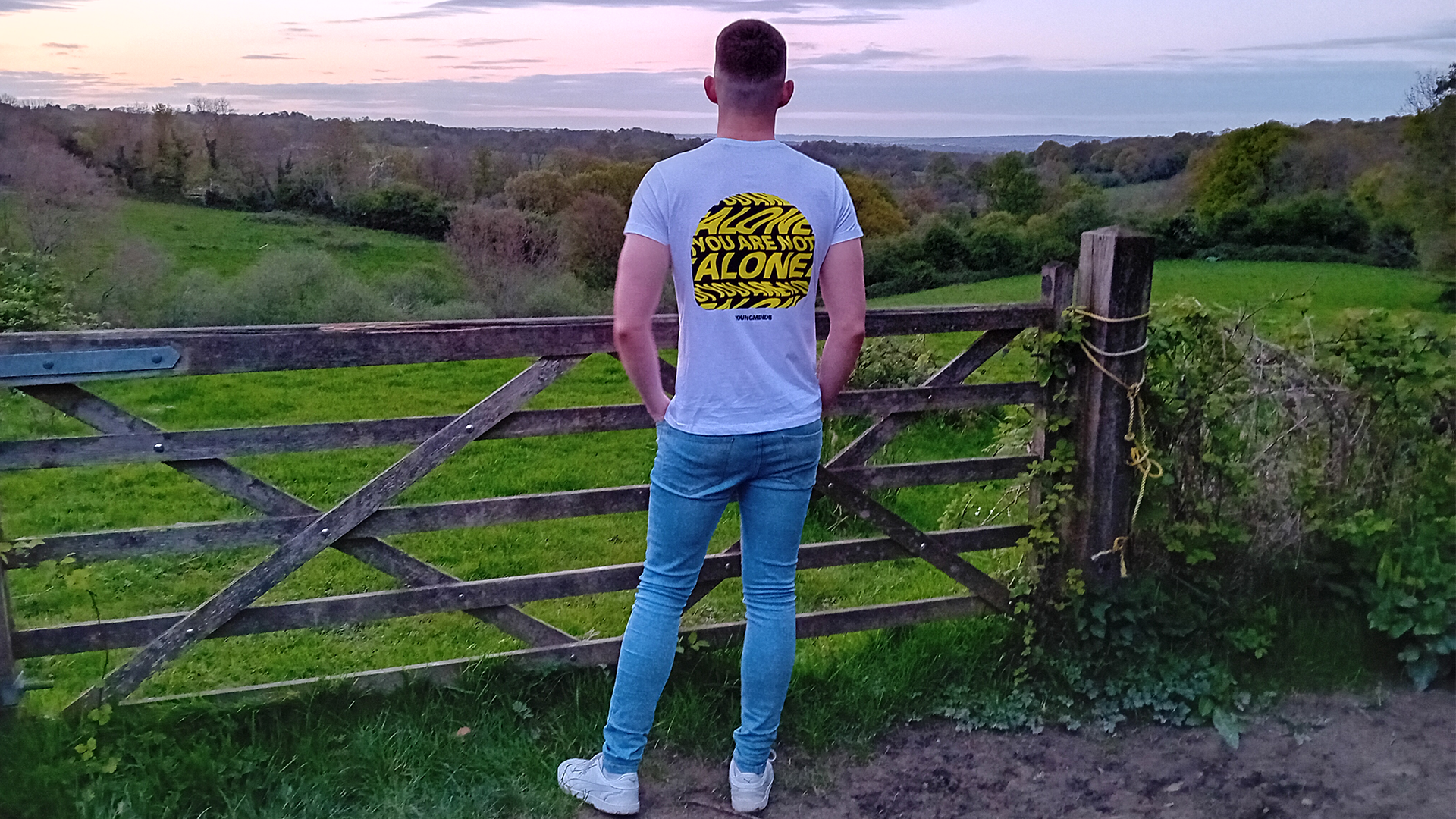 A person stands with their back to the camera, wearing a YoungMinds t-shirt. They are looking out over the fields in the countryside.