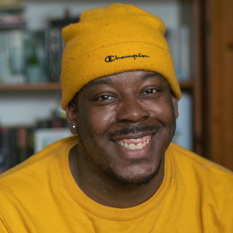 A picture of Femi wearing a yellow hat and yellow top and smiling at the camera.