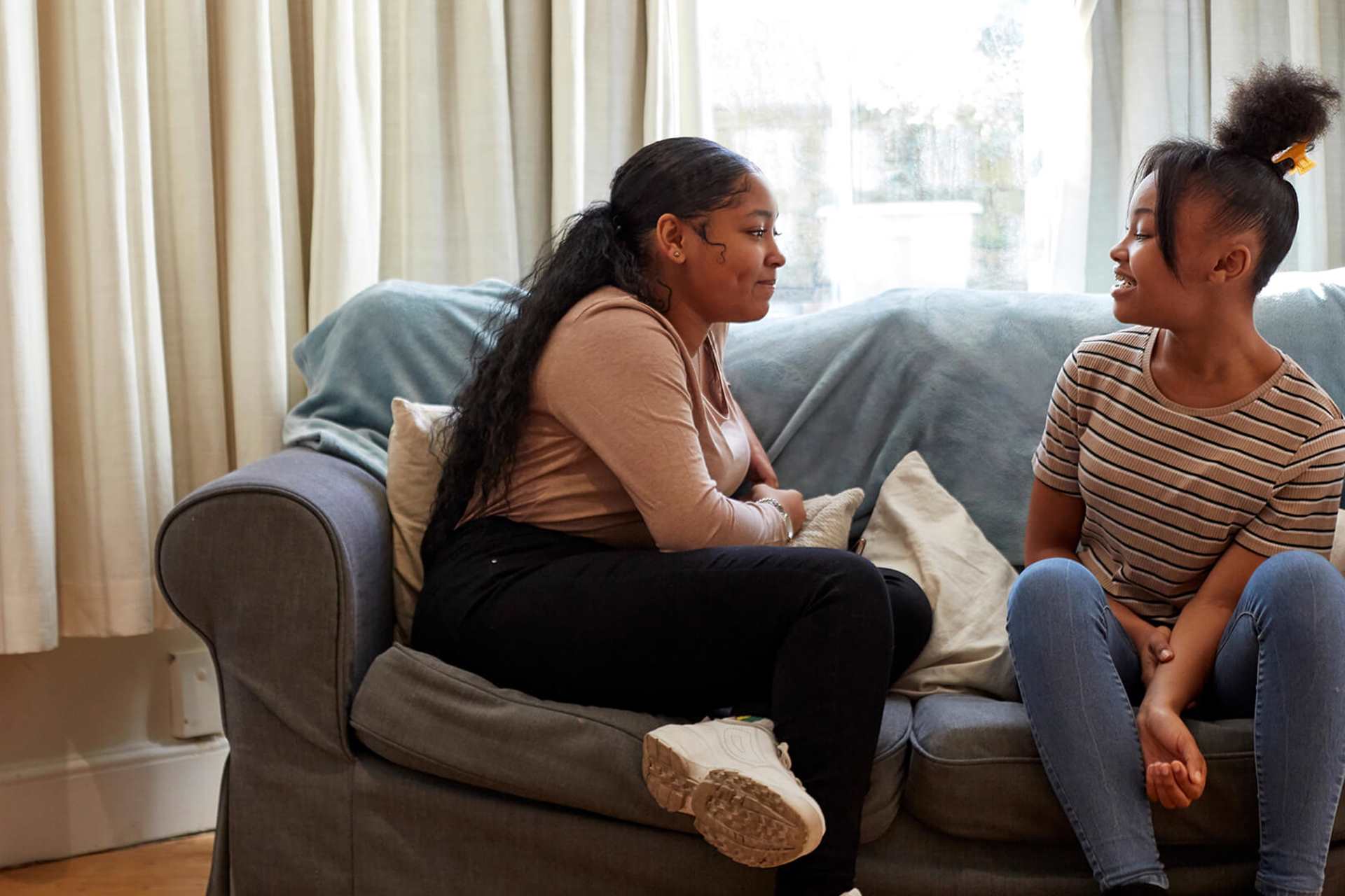 Two girls sitting on a blue sofa. They are looking at each other and talking.