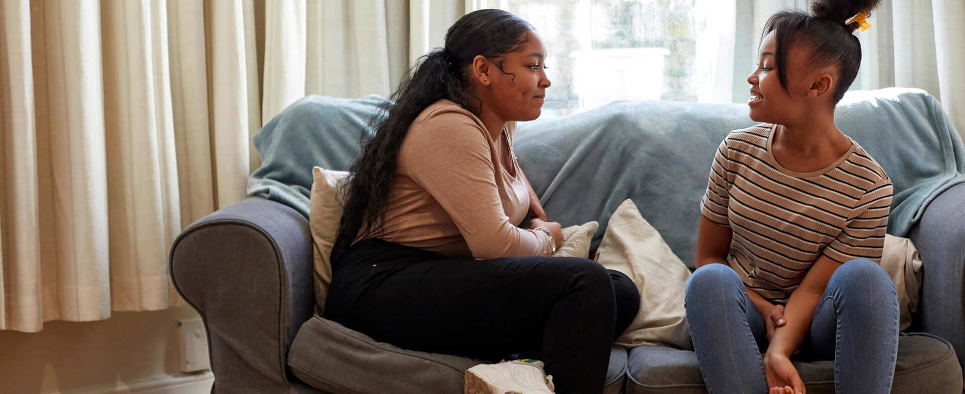 Two girls sitting on a blue sofa. They are looking at each other and talking.