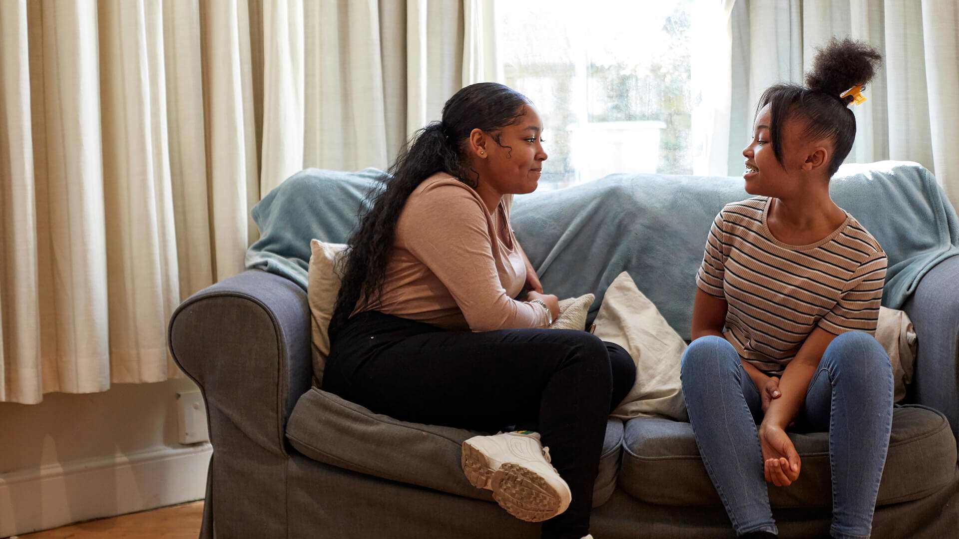 Two girls sitting on a blue sofa. They are looking at each other and talking.