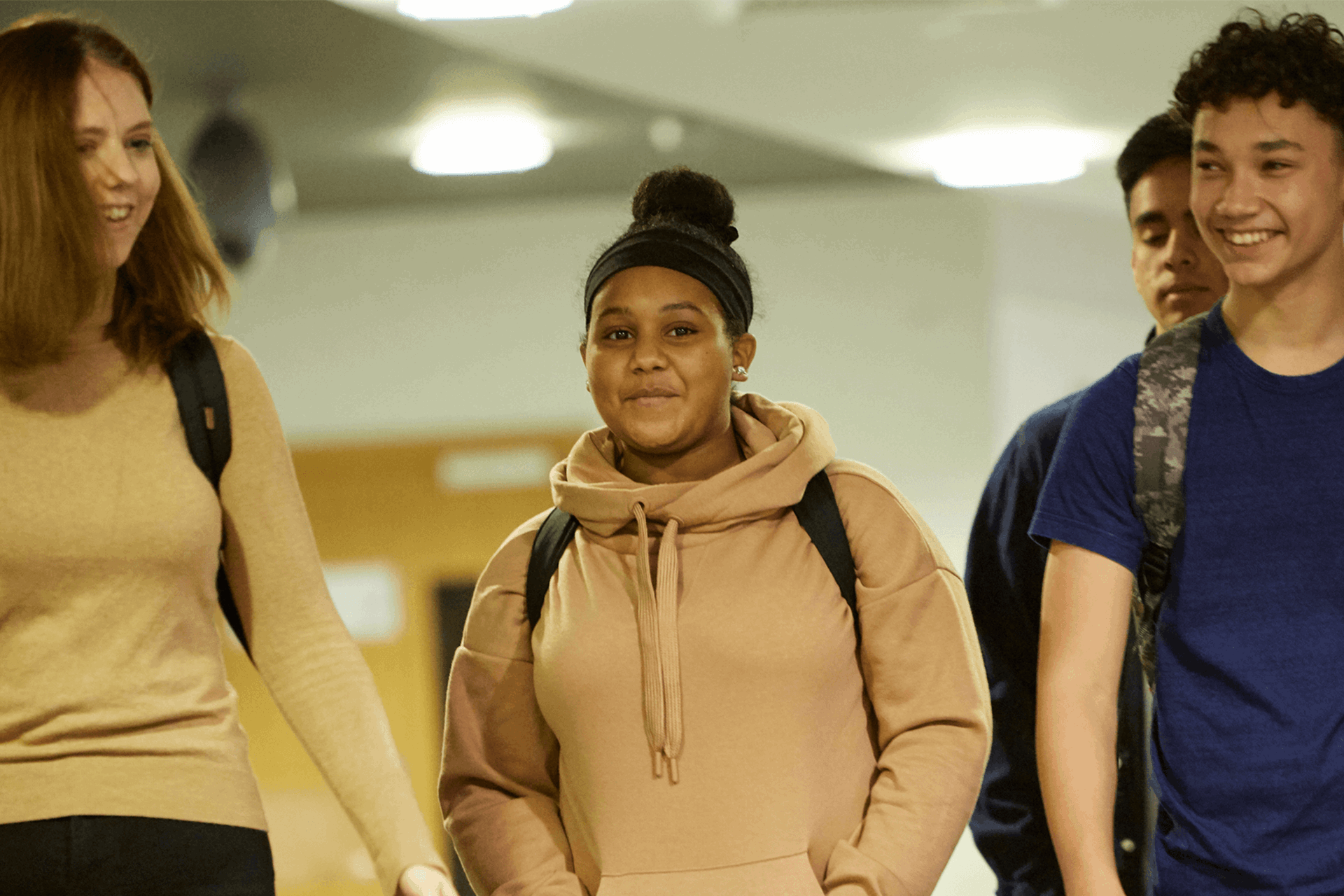 Four students wearing backpacks and smiling while walking down a school hallway together.