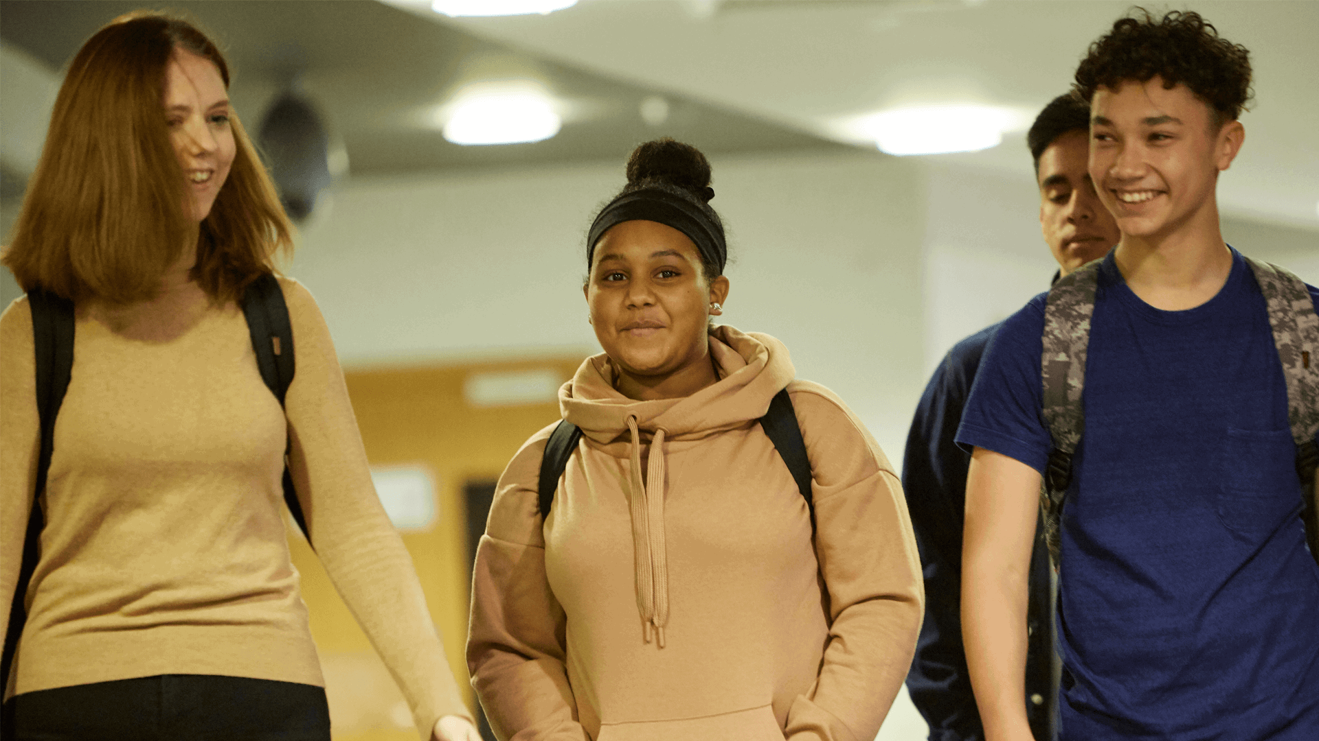 Four students wearing backpacks and smiling while walking down a school hallway together.