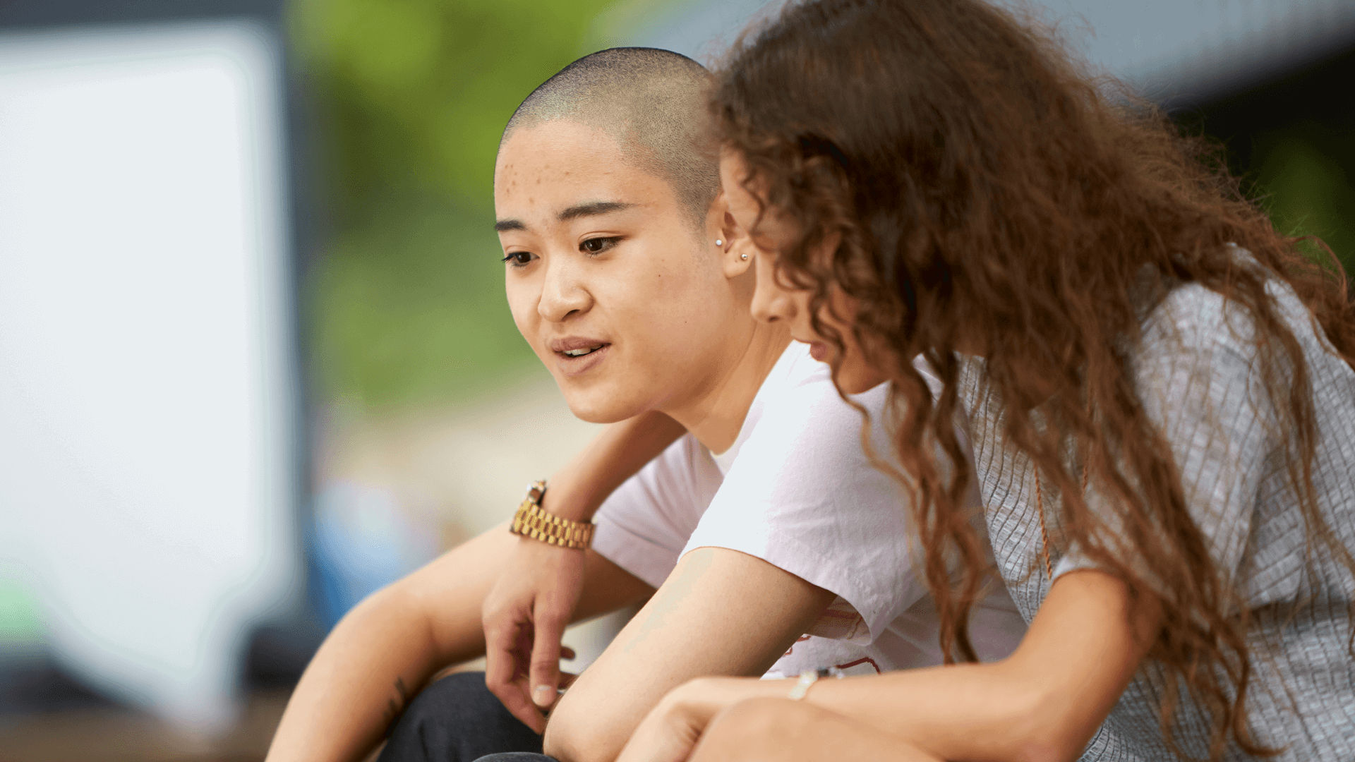 A girl with curly hair has her arm over her girlfriend's shoulder and talks to her while they sit in a park.