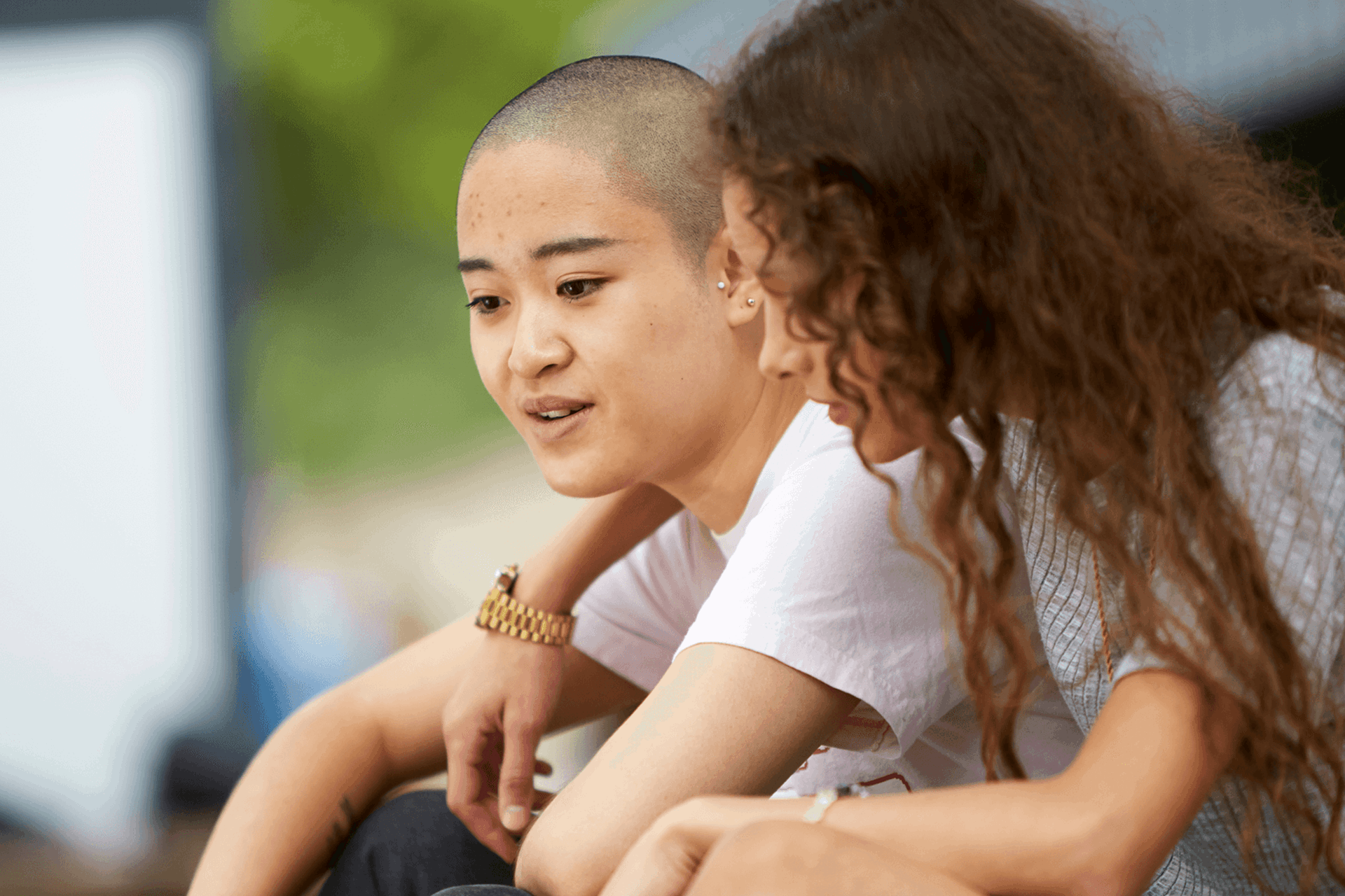 A girl with curly hair has her arm over her girlfriend's shoulder and talks to her while they sit in a park.