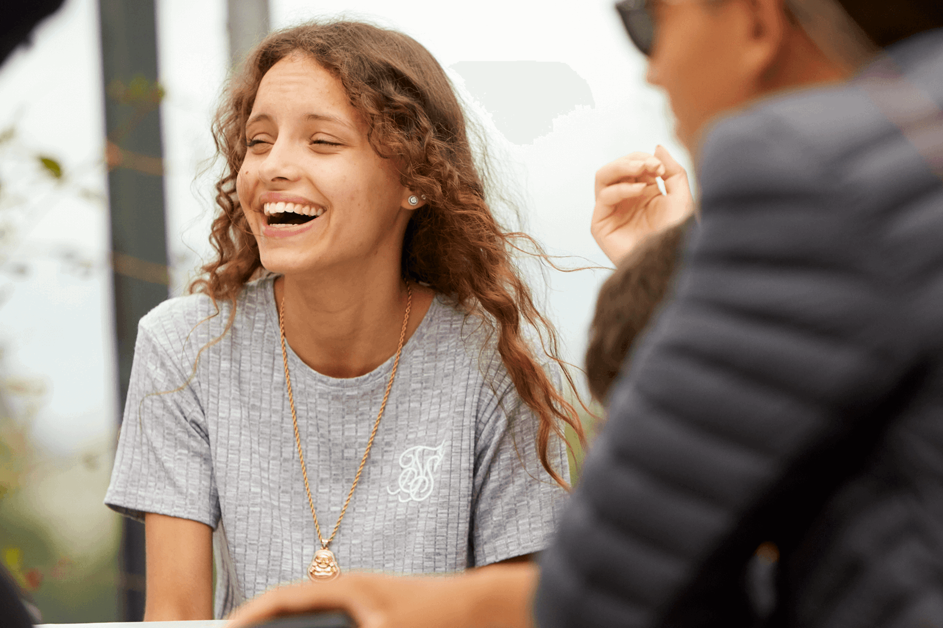 A young woman with long, curly hair wearing a grey T-shirt. She is sitting with her friends and laughing.