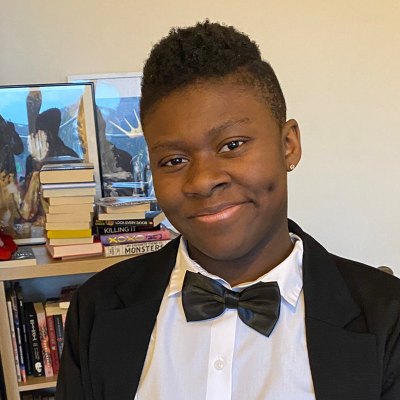 Simi is a Black young person and they are wearing a Black tuxedo with bow tie whilst sitting in their wheelchair and smiling.