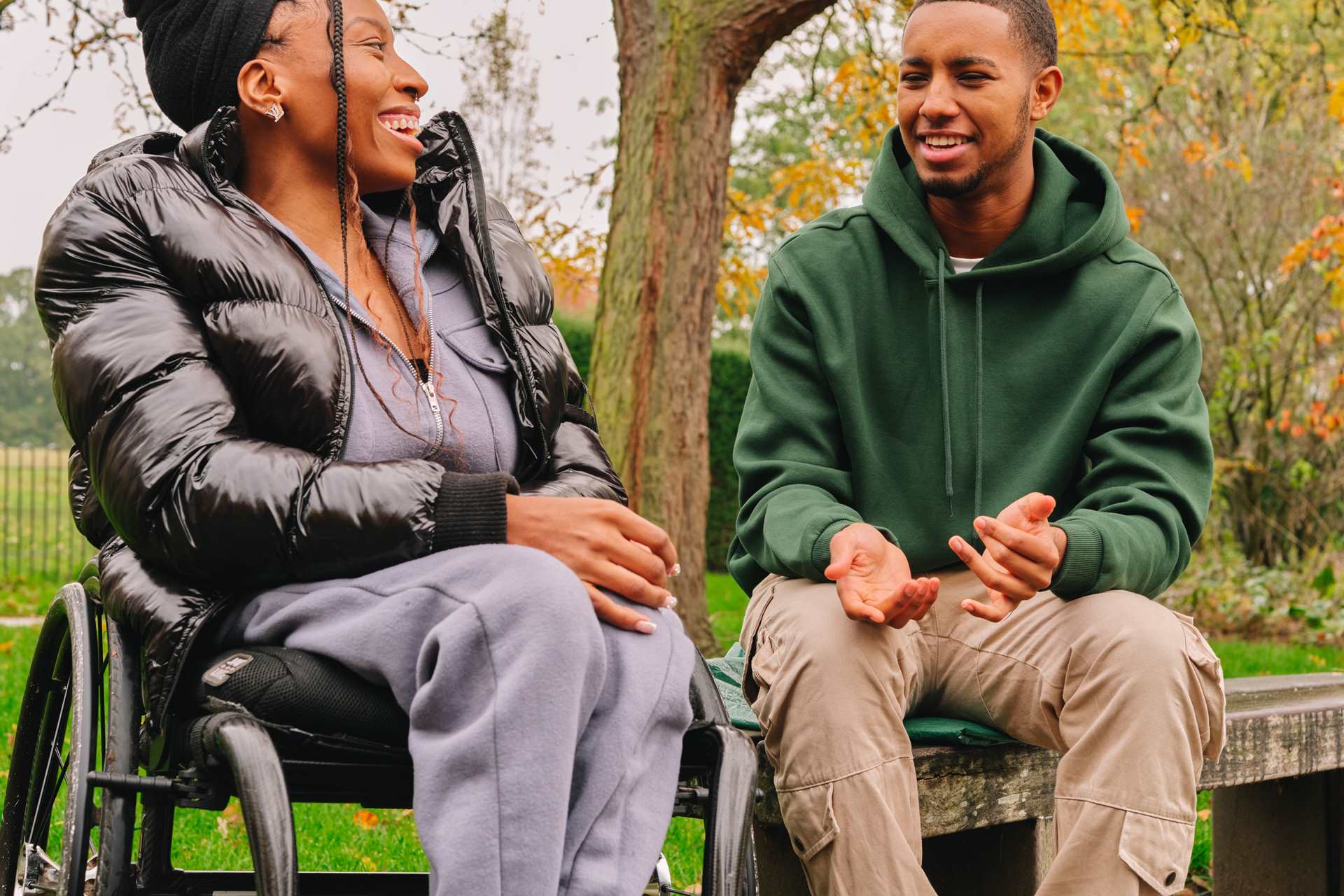 A young Black woman in a wheelchair and a young Black man on a bench. They are talking and laughing together.
