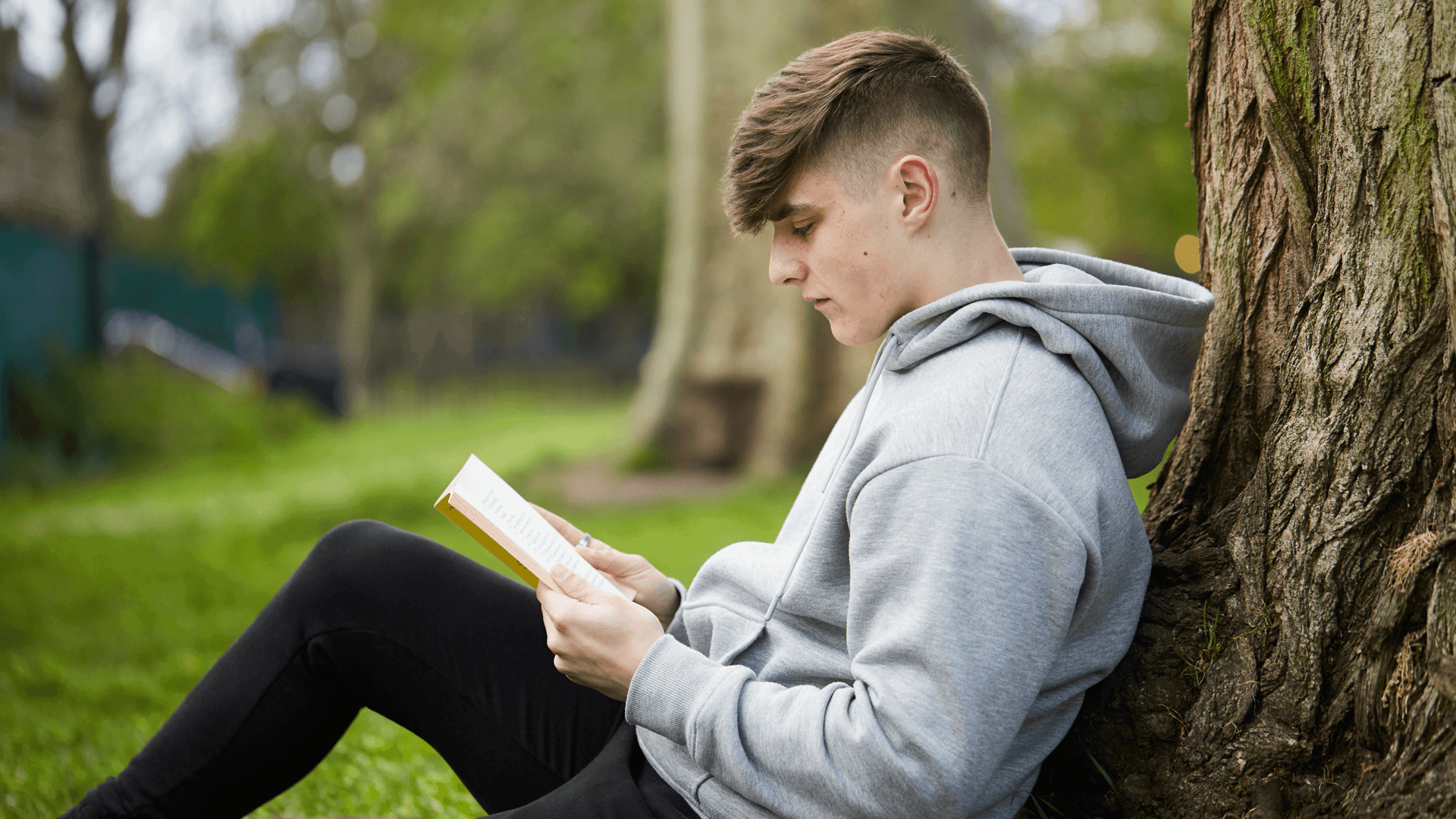 A young person sits against a tree in the park while reading a book.