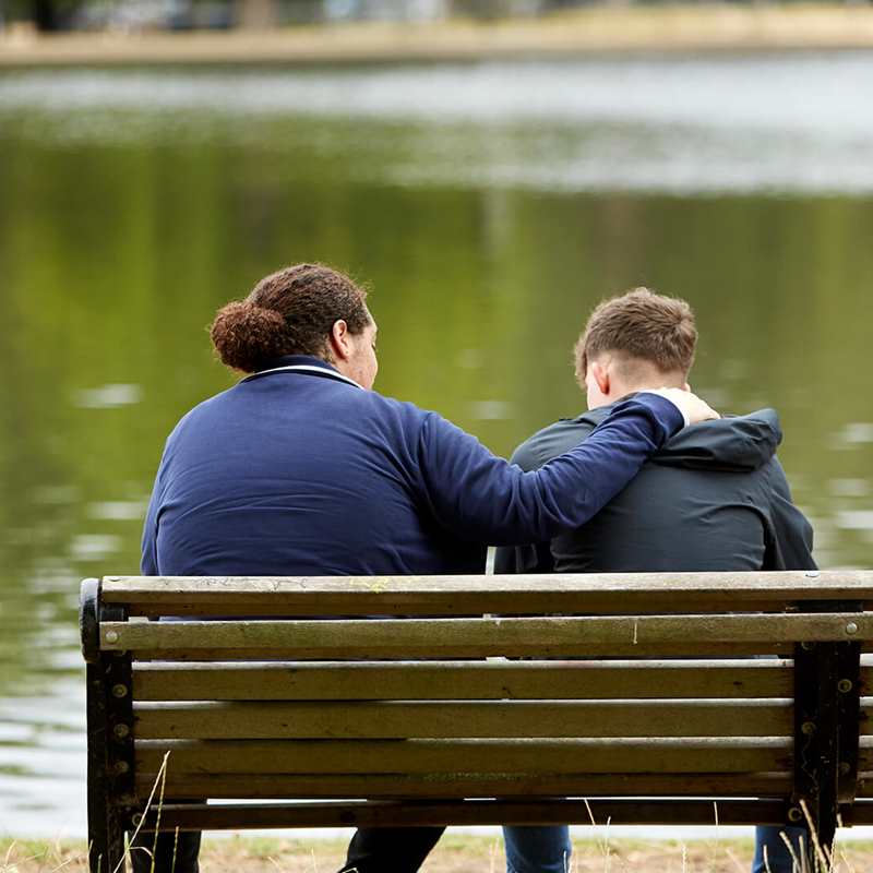 back-shot-of-two-boys-wearing-jackets-comforting-each-other-while-sitting-on-a-park-bench-in-front-of-a-lake