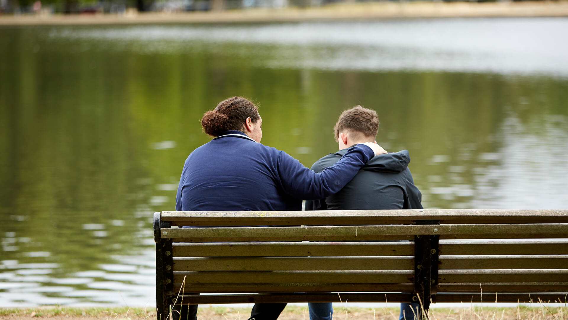 Two young men sit on a park bench overlooking a lake. One has his arm on the other's shoulder to comfort him.