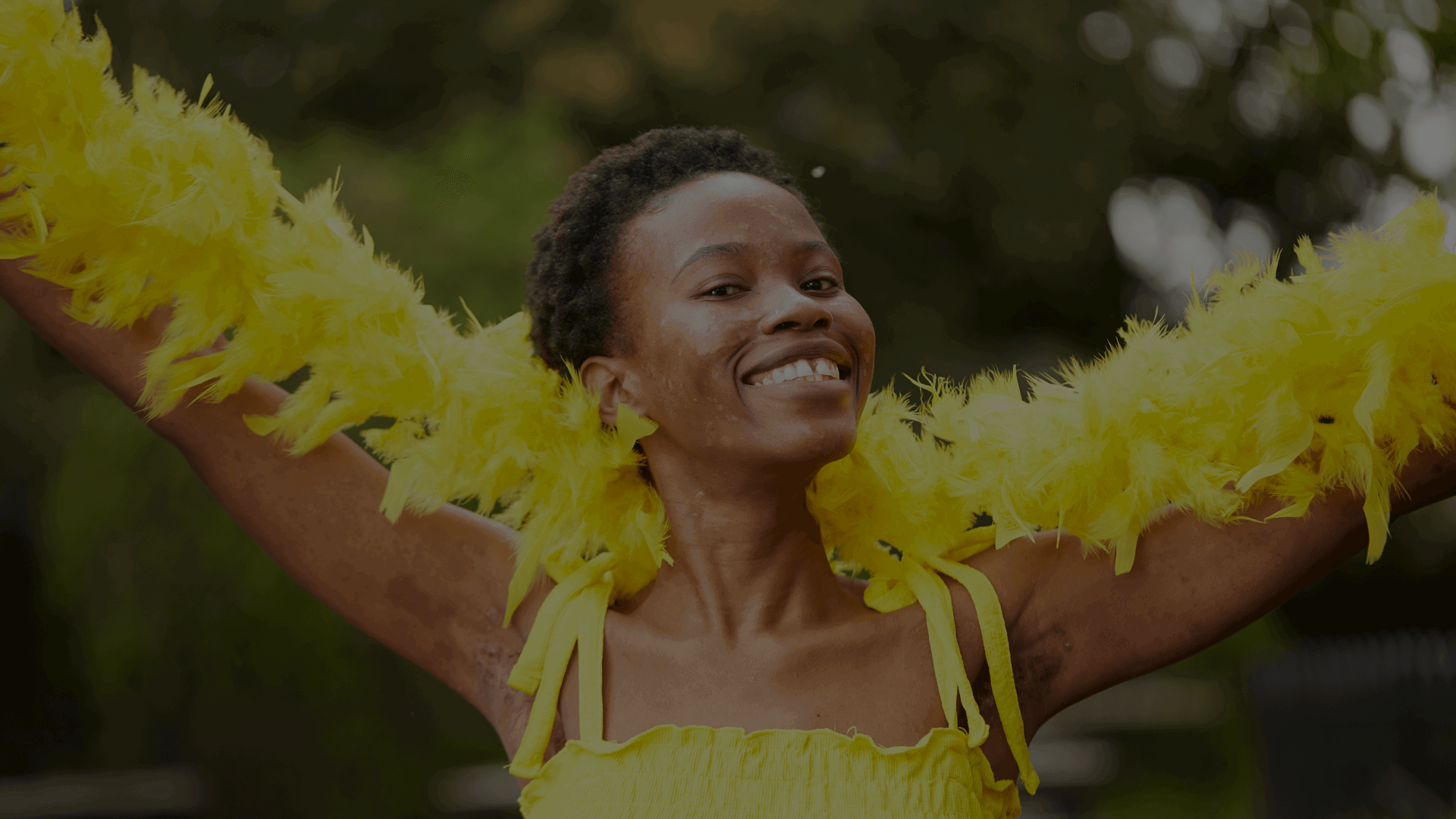 A young person has their arms stretched out while holding a feather boa. They are wearing a yellow top, looking towards the camera and smiling.