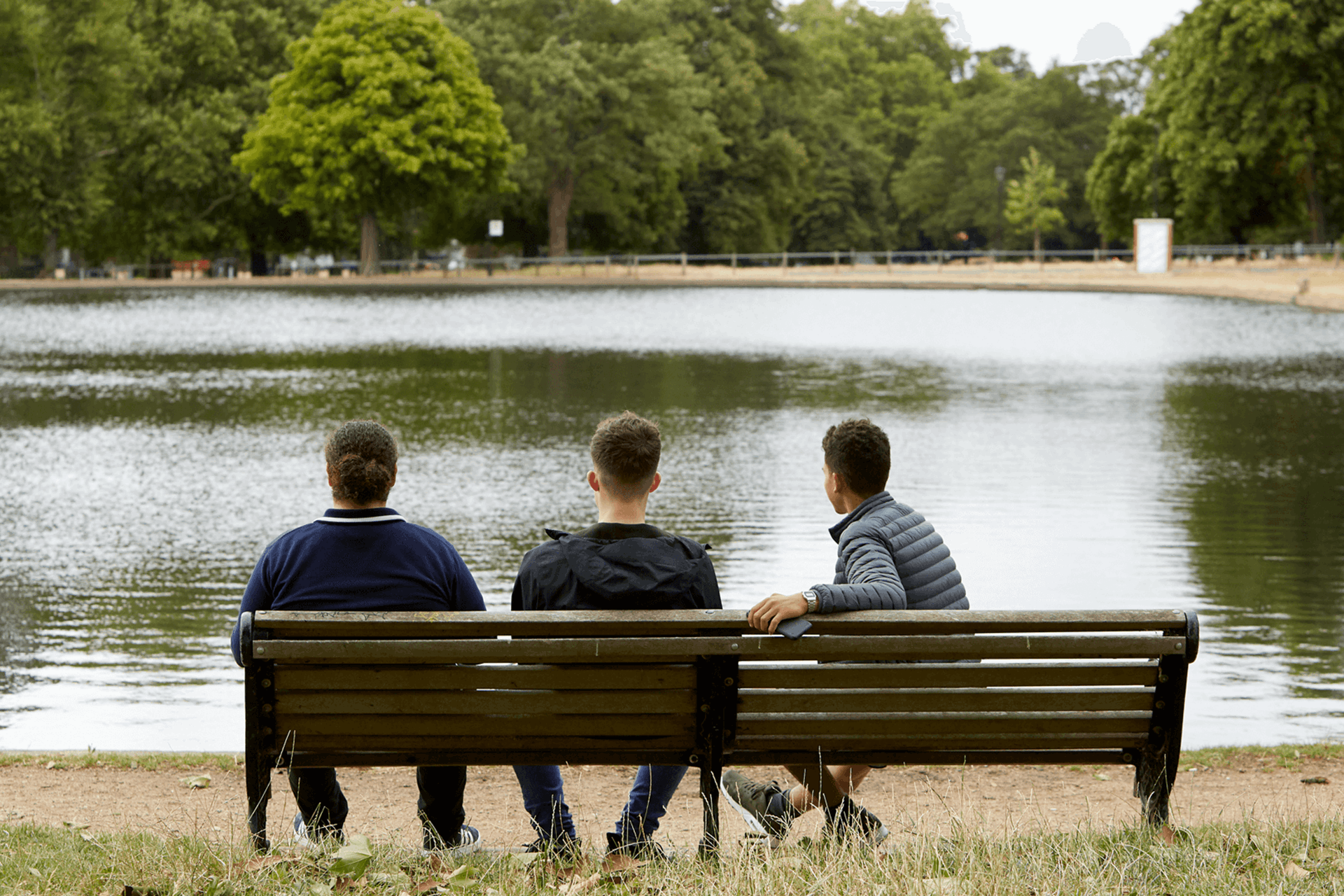 wide shot of three boys sitting on a bench looking over the lake in the park