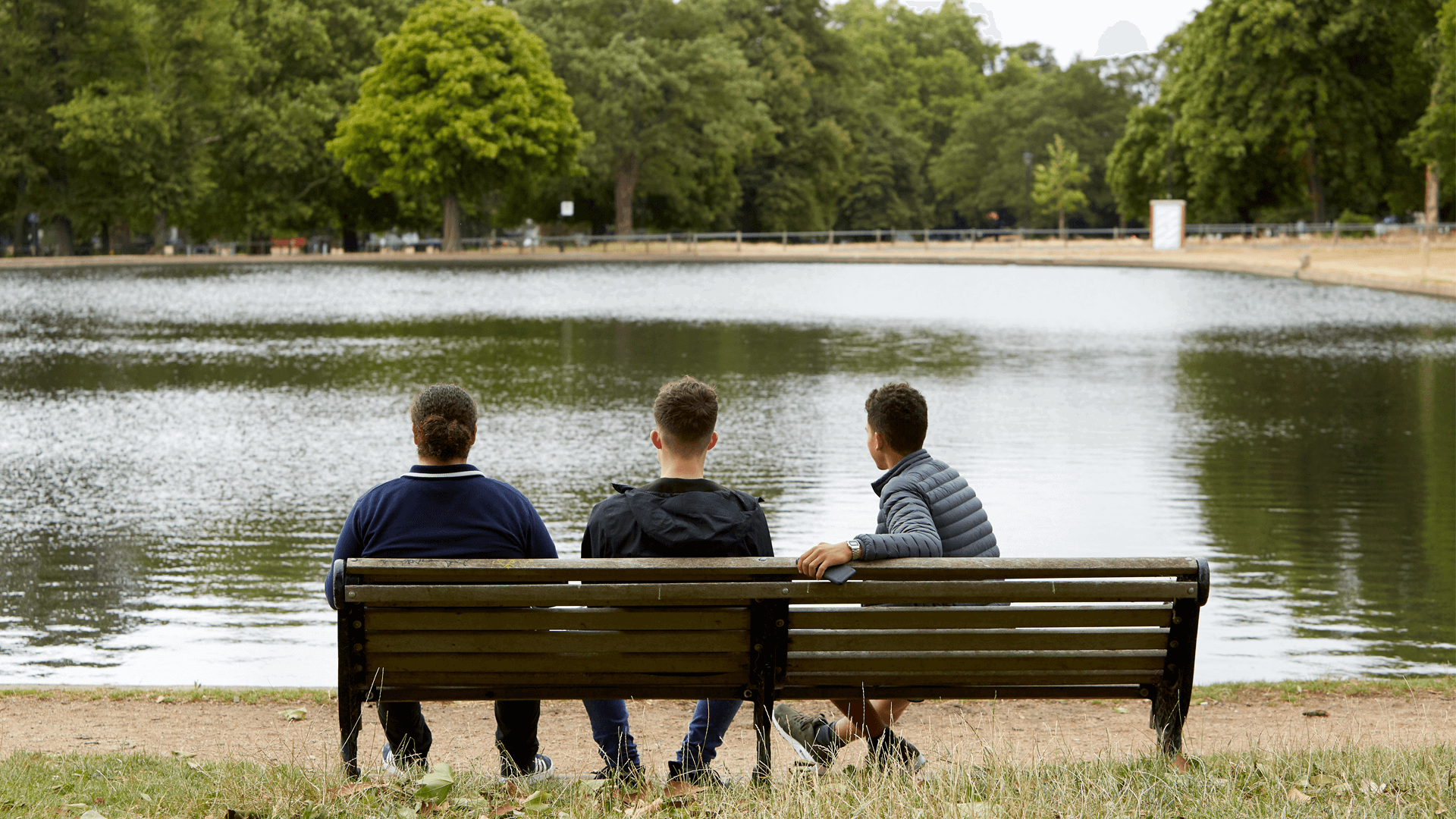 wide shot of three boys sitting on a bench looking over the lake in the park