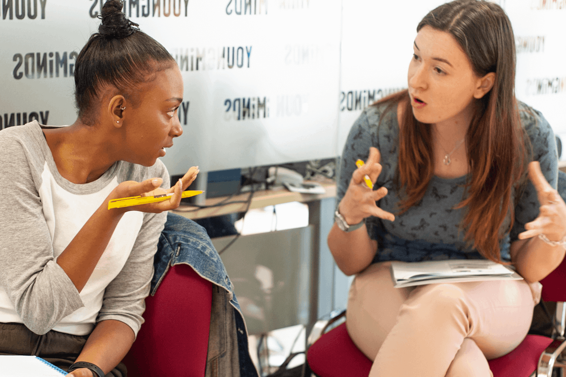 two moms are having a discussion over an activity while sitting on chairs inside a conference room