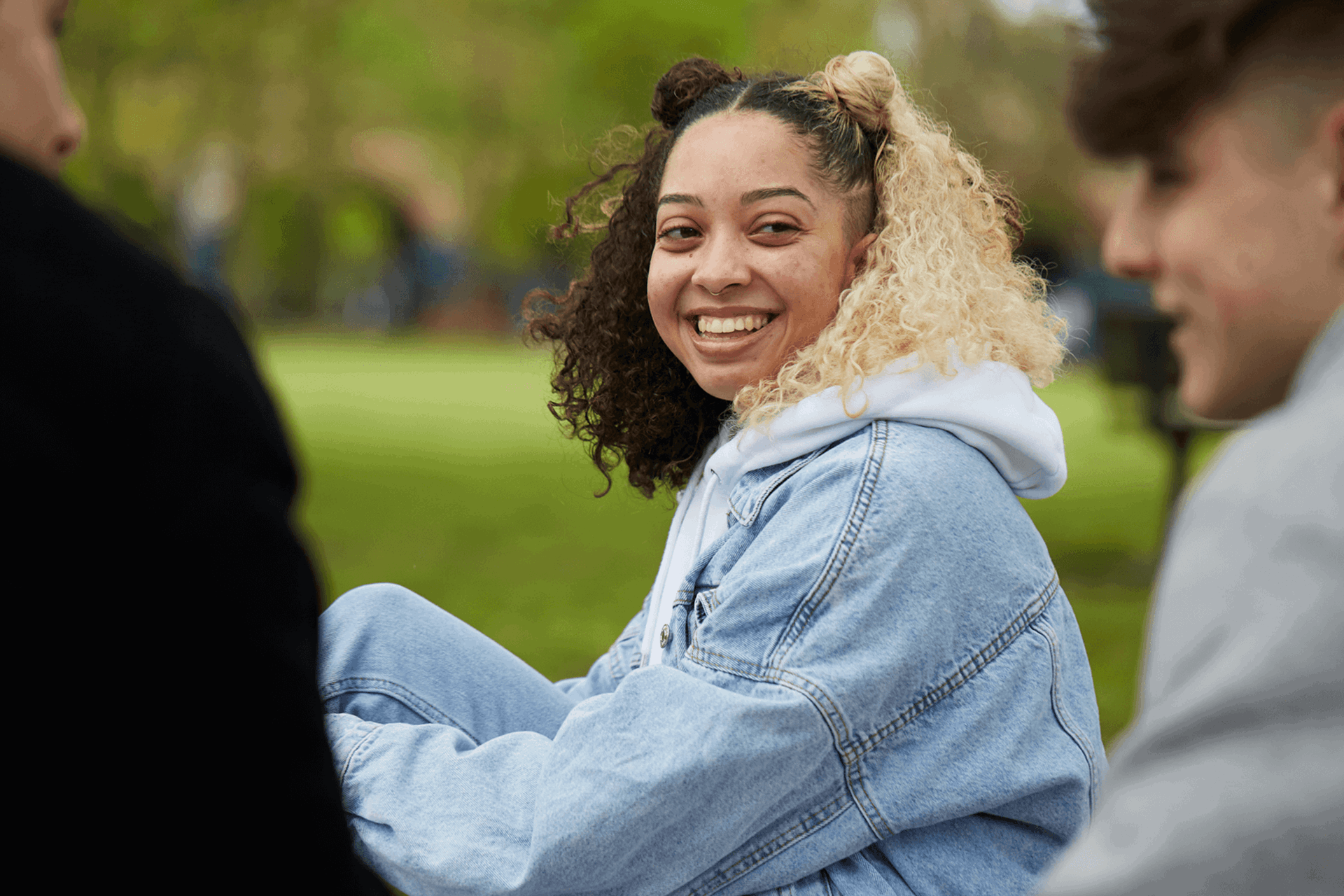 A young woman with curly hair wearing a blue denim jacket is smiling and talking with two young men in the park.
