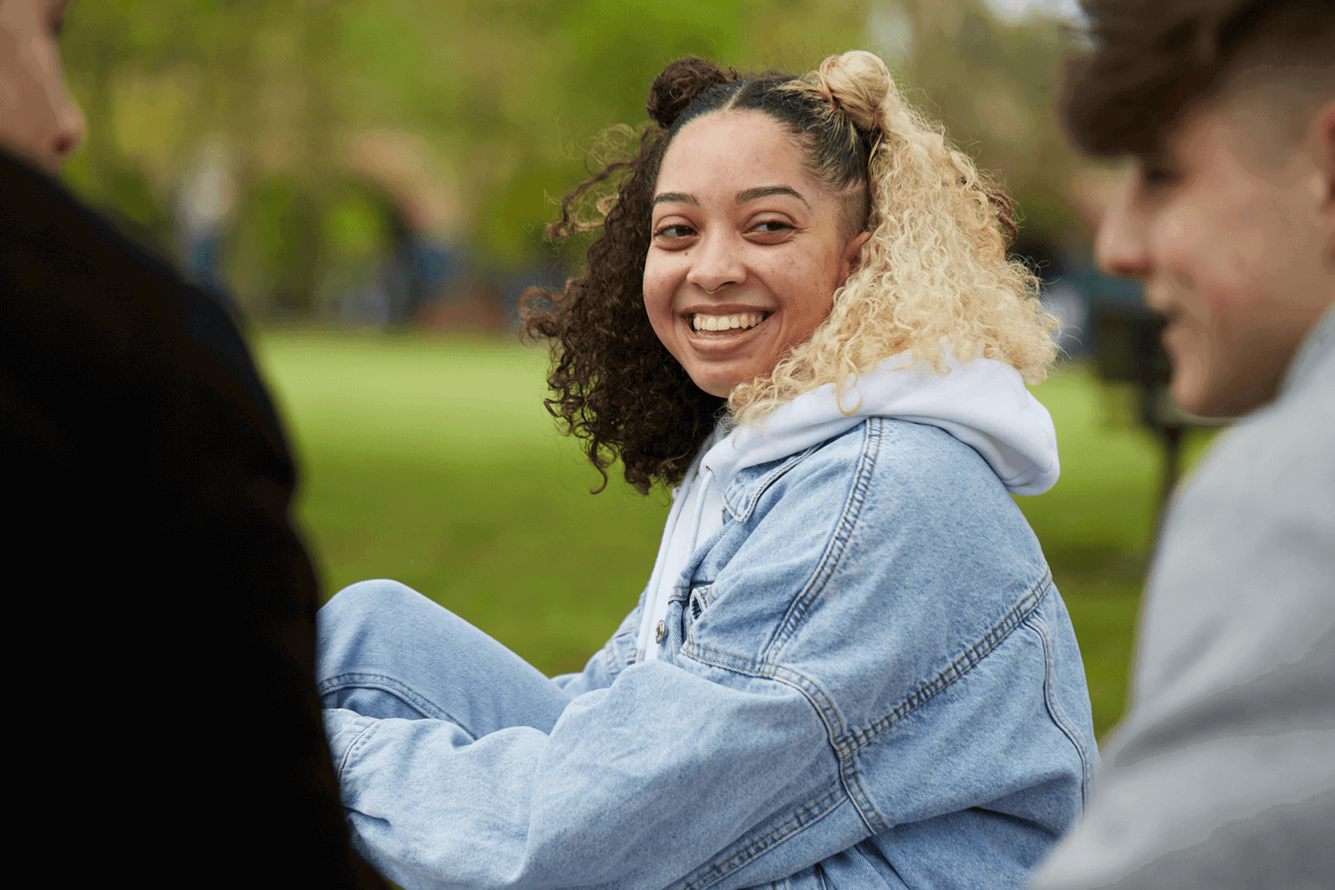 A young woman with curly hair wearing a blue denim jacket is smiling and talking with two young men in the park.