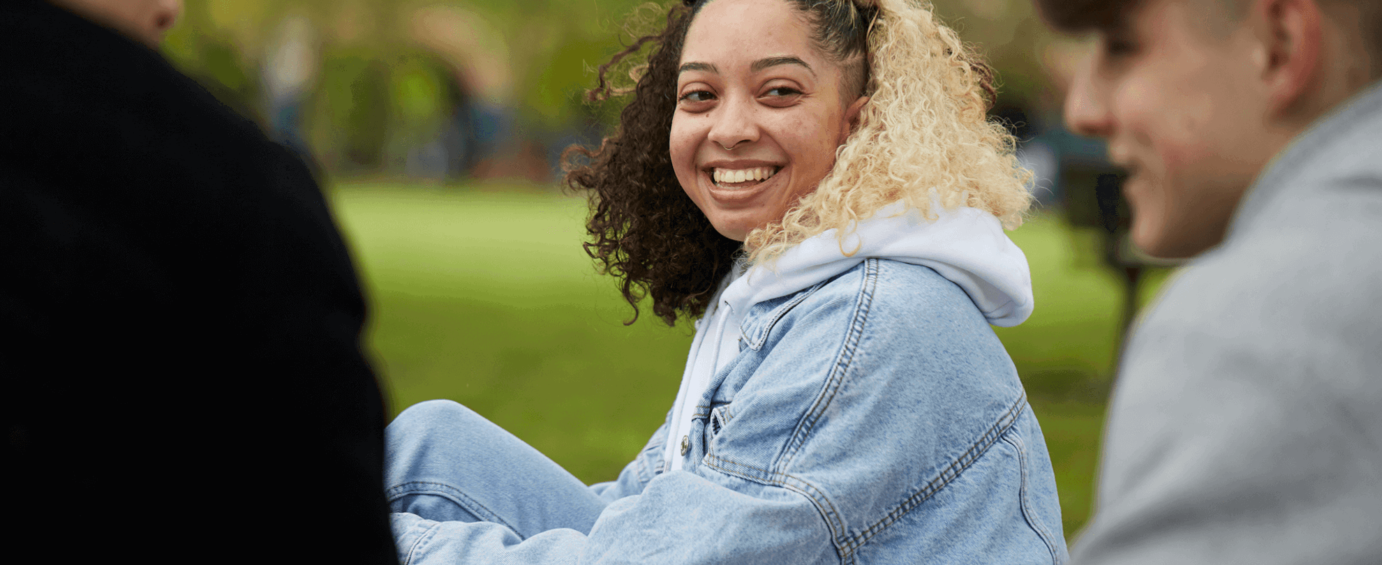 A young woman with curly hair wearing a blue denim jacket is smiling and talking with two young men in the park.