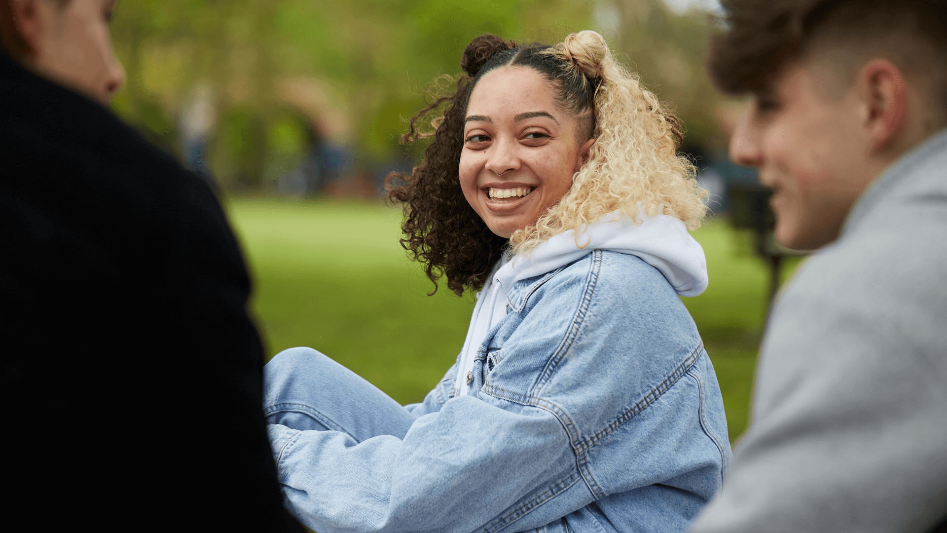 A young woman with curly hair wearing a blue denim jacket is smiling and talking with two young men in the park.