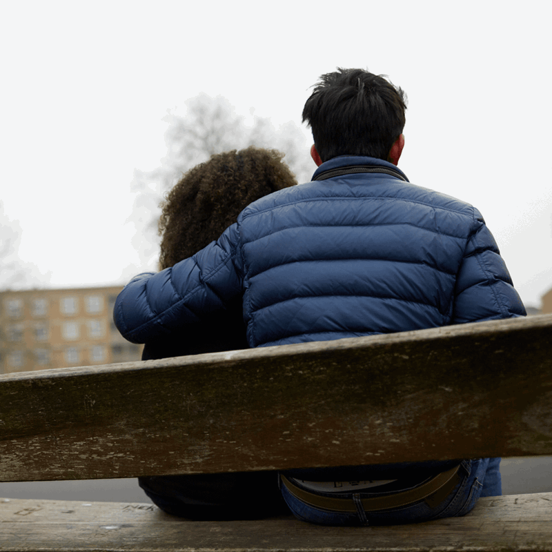backshot-of-a-boy-with-arms-around-a-girl-while-sitting-on-a-bench-in-a-park-with-a-gloomy-sky-on-background