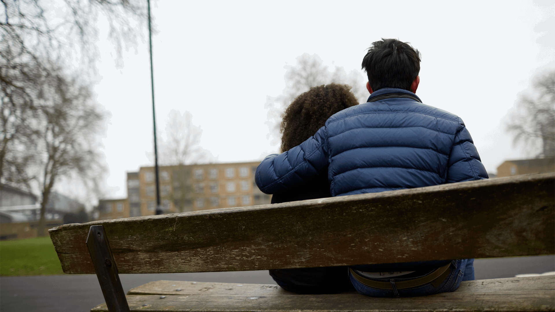 Two young people sitting together on a bench. One has their arm around the other.