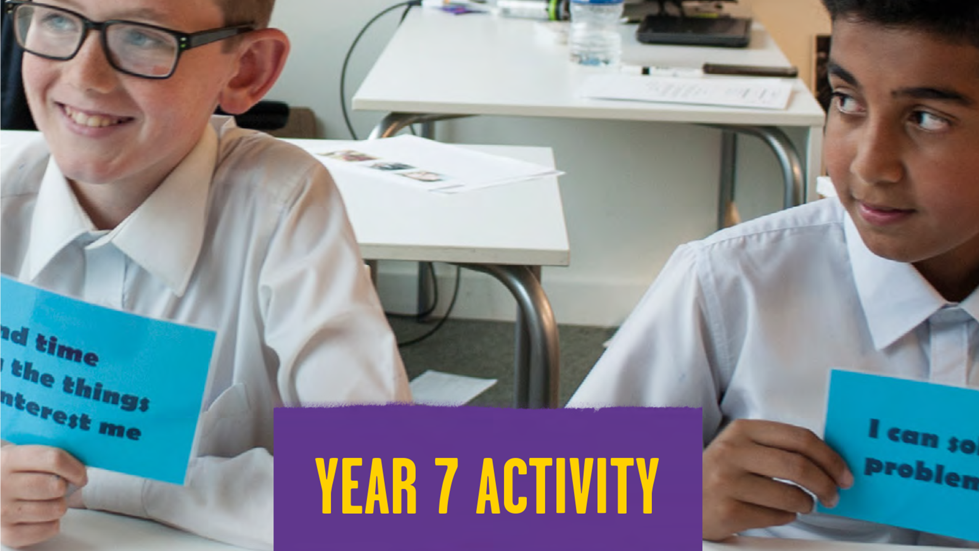 Two students sit next to each at a desk in a classroom holding blue cards with different statements on. In the middle of the students, there's a purple square with yellow writing. It says 'year 7 activity'.