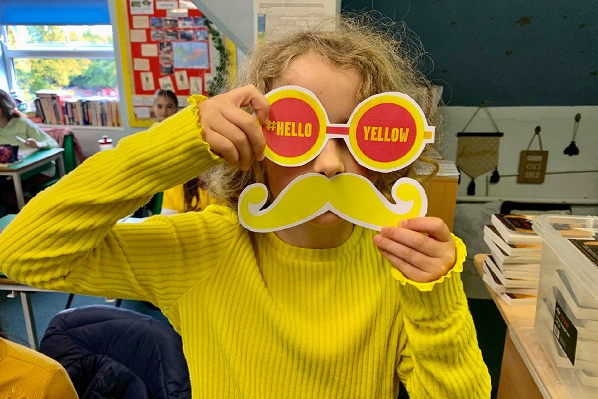 A young person stands holding glasses that have helloyellow written across them and a yellow moustache. The young person is wearing a bright yellow jumper.