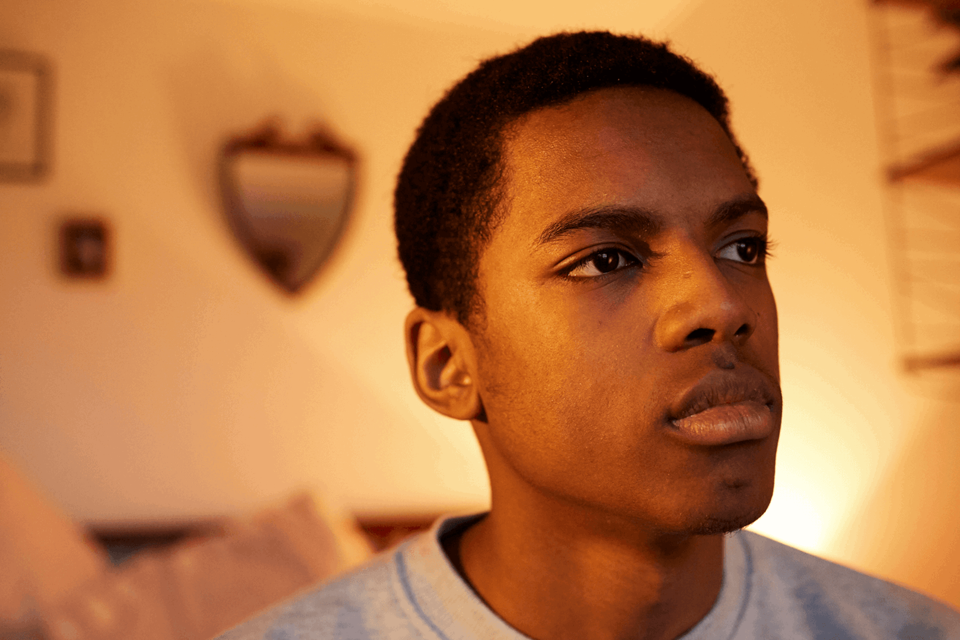 A boy sitting alone on the end of his bed and appearing deep in thought.