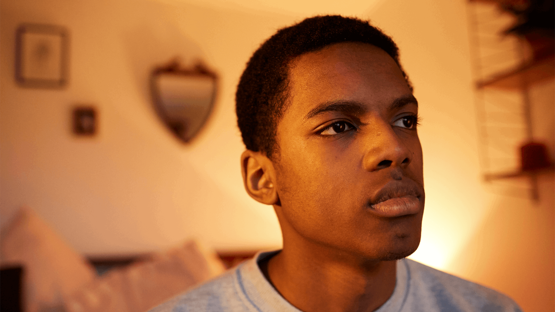 A boy sitting alone on the end of his bed and appearing deep in thought.