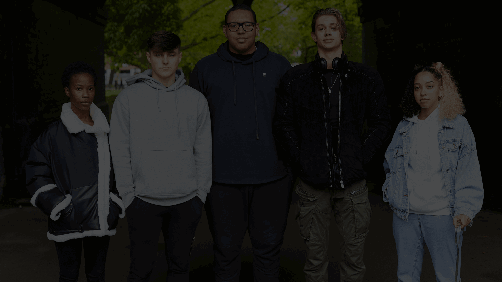 Five young people all stand next each other, looking serious at the camera. They are standing underneath a bridge with trees in the background.