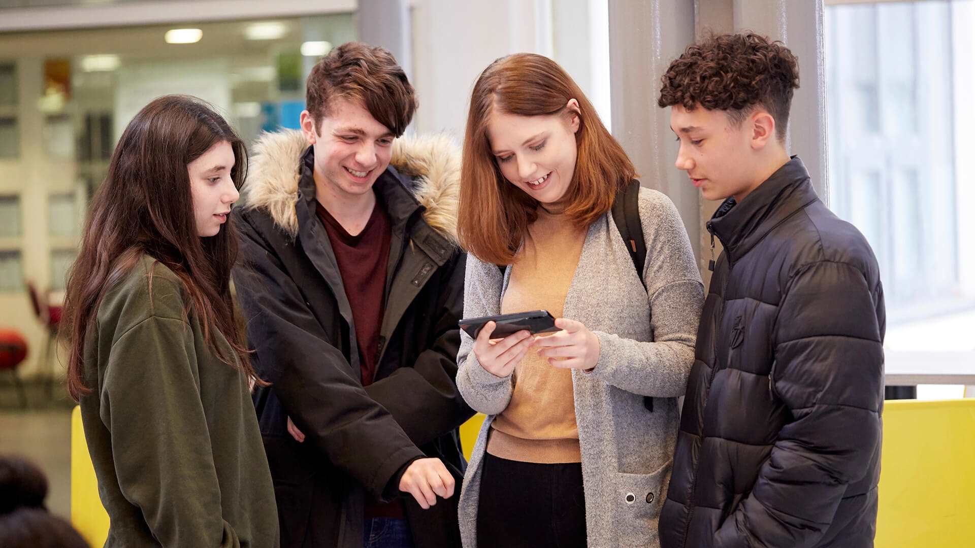 Four young people huddle round together, smiling and laughing, looking at a phone that the person in the middle is holding. They stand inside a campus building.