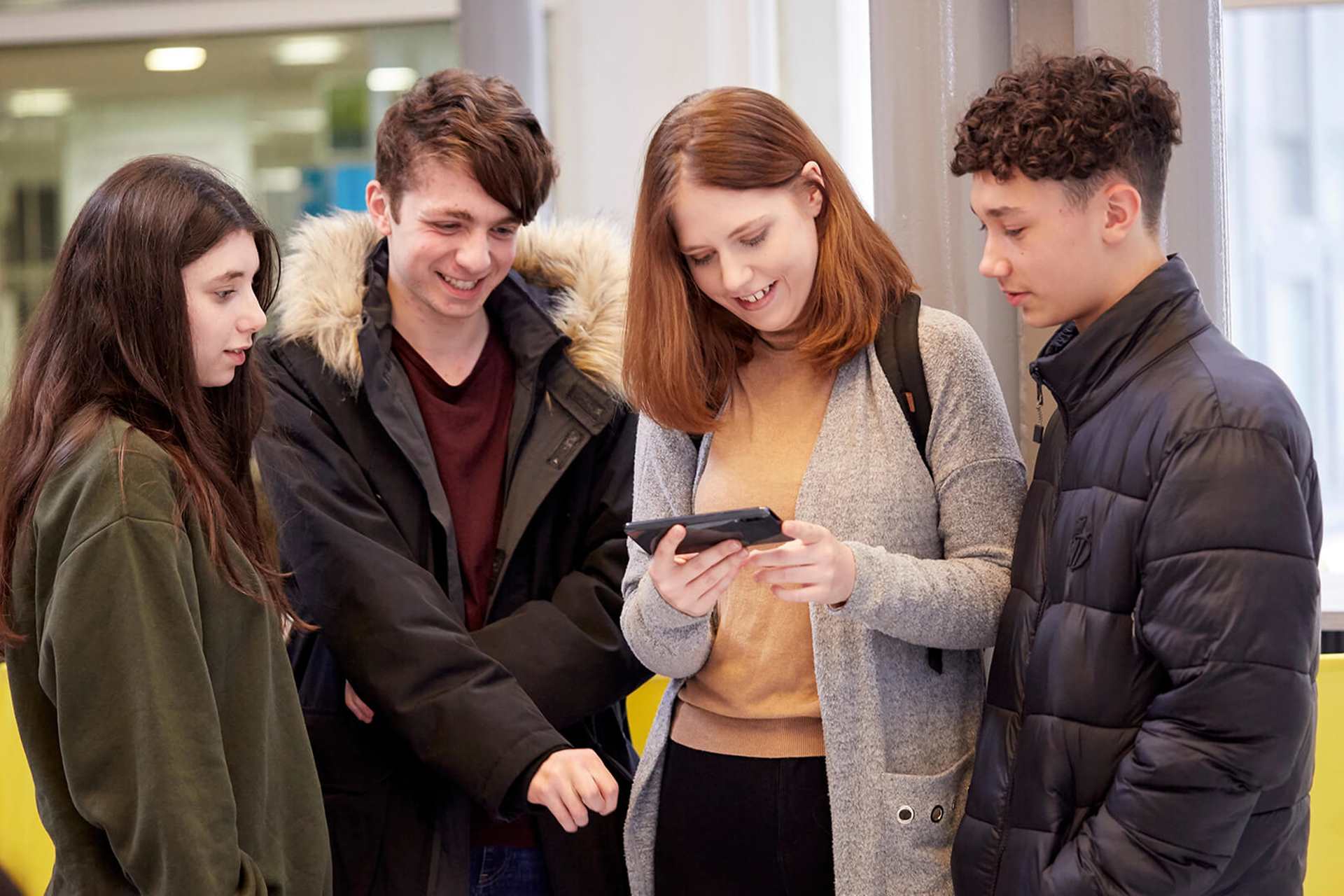 Four young people huddle round together, smiling and laughing, looking at a phone that the person in the middle is holding. They stand inside a campus building.