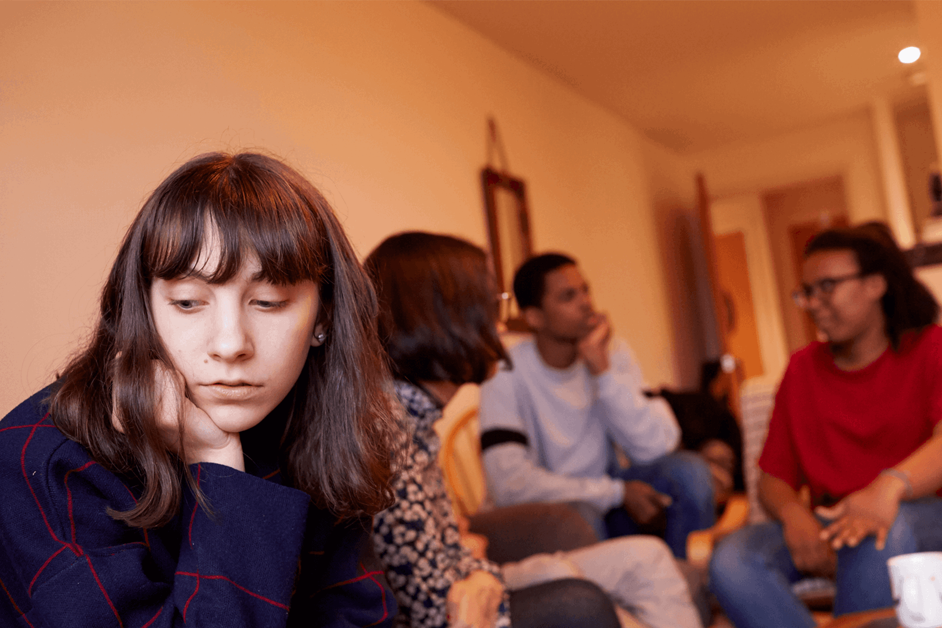 A girl sitting in a living room with her friends. She is looking away as they talk behind her and seems left out of the conversation.