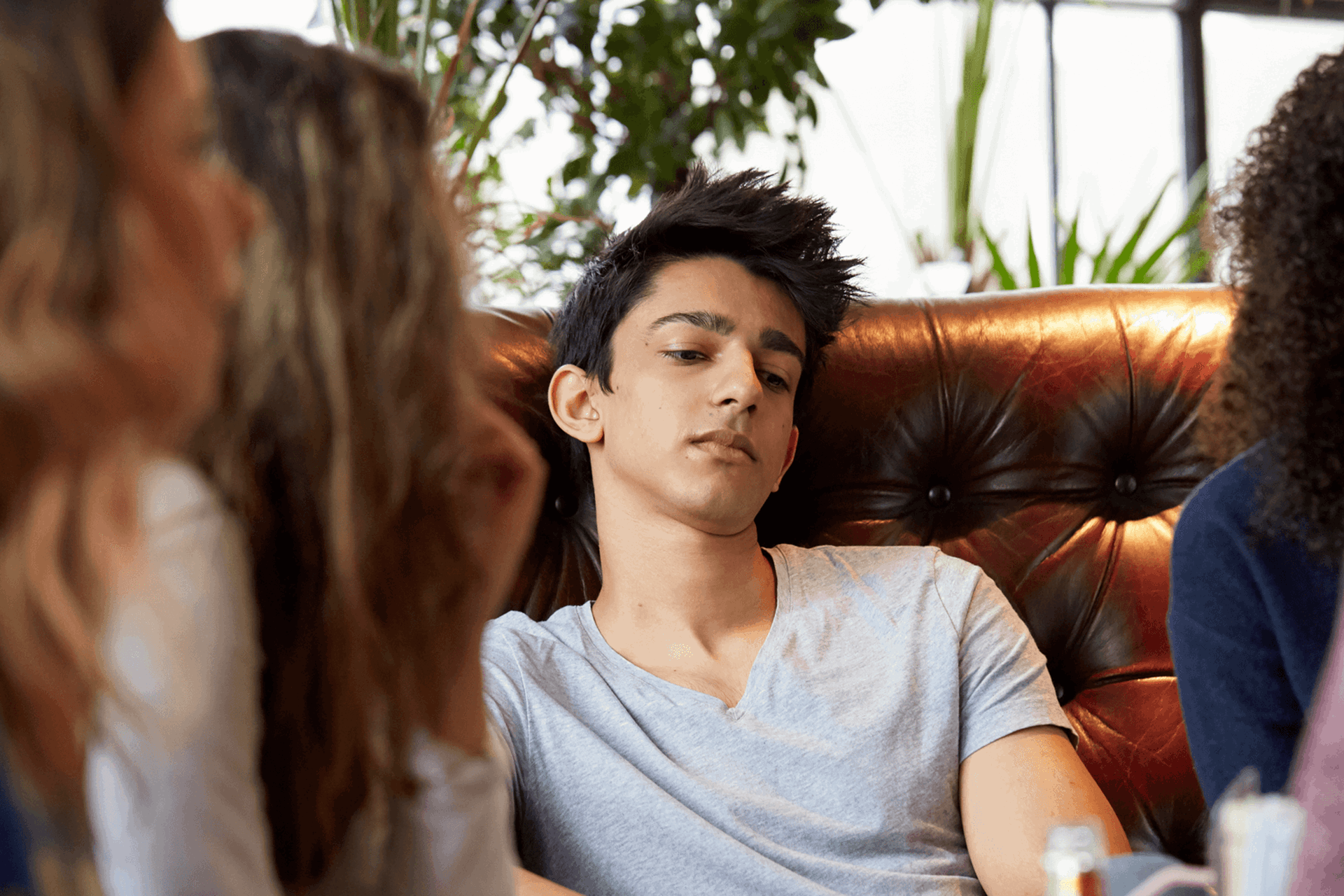 a boy wearing white shirt looking lost in thoughts as he sit on a couch surrounded by friends chatting