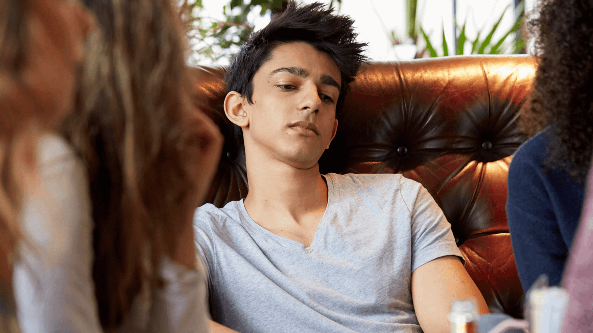 a boy wearing white shirt looking lost in thoughts as he sit on a couch surrounded by friends chatting