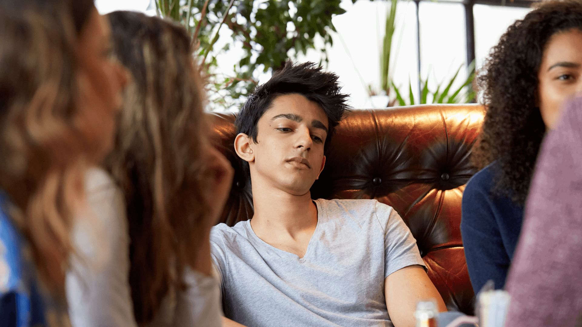 a boy wearing white shirt looking lost in thoughts as he sit on a couch surrounded by friends chatting