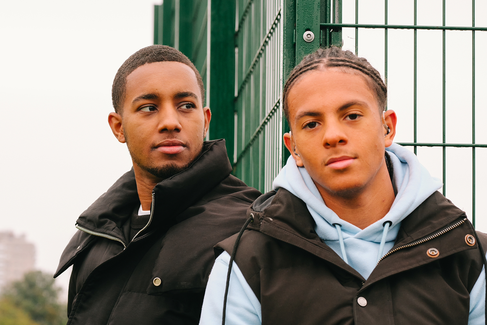 A young Black man sitting in the park with a Black teenage boy wearing a hearing aid. They are both looking very serious.