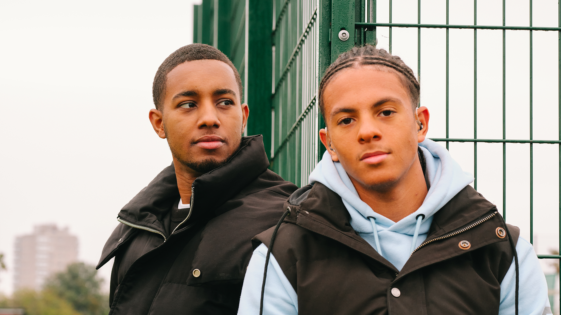 A young Black man sitting in the park with a Black teenage boy wearing a hearing aid. They are both looking very serious.