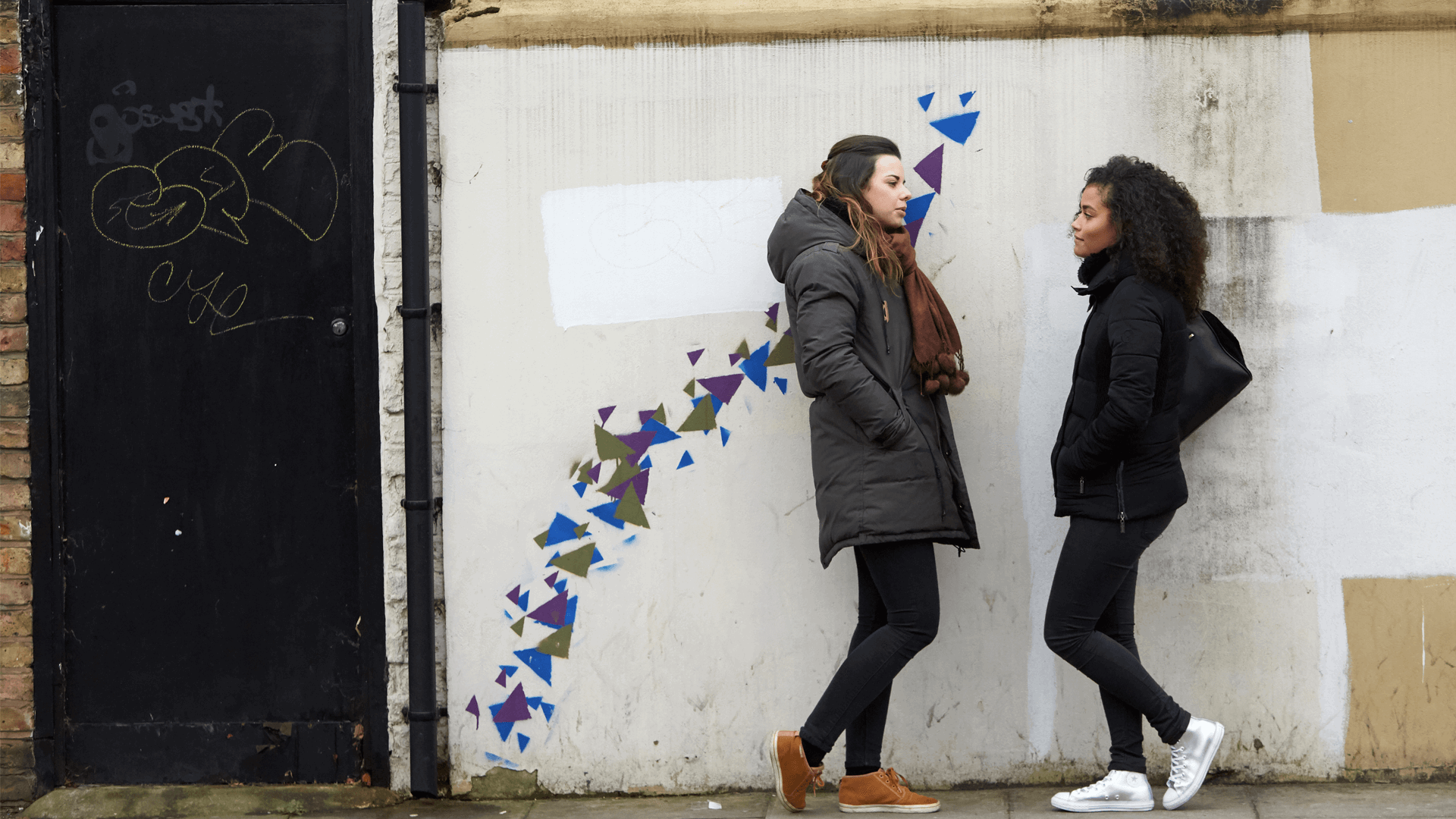Two girls wearing black jackets are talking while standing against a wall decorated with blue, purple and green graffiti patterns.