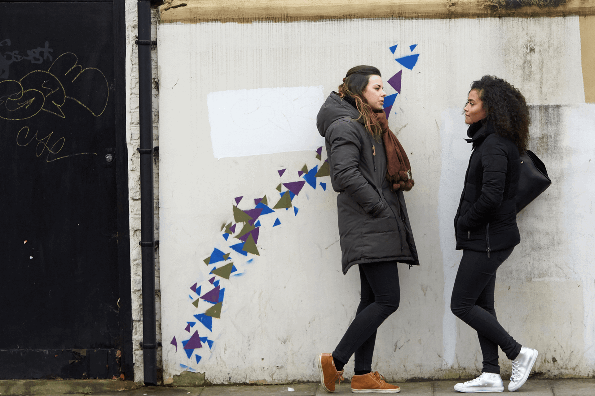 Two girls wearing black jackets are talking while standing against a wall decorated with blue, purple and green graffiti patterns.
