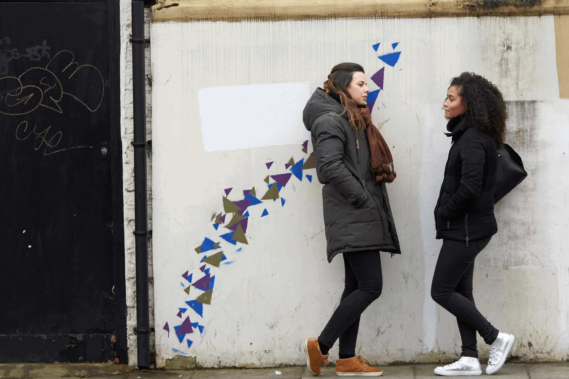 Two girls wearing black jackets are talking while standing against a wall decorated with blue, purple and green graffiti patterns.