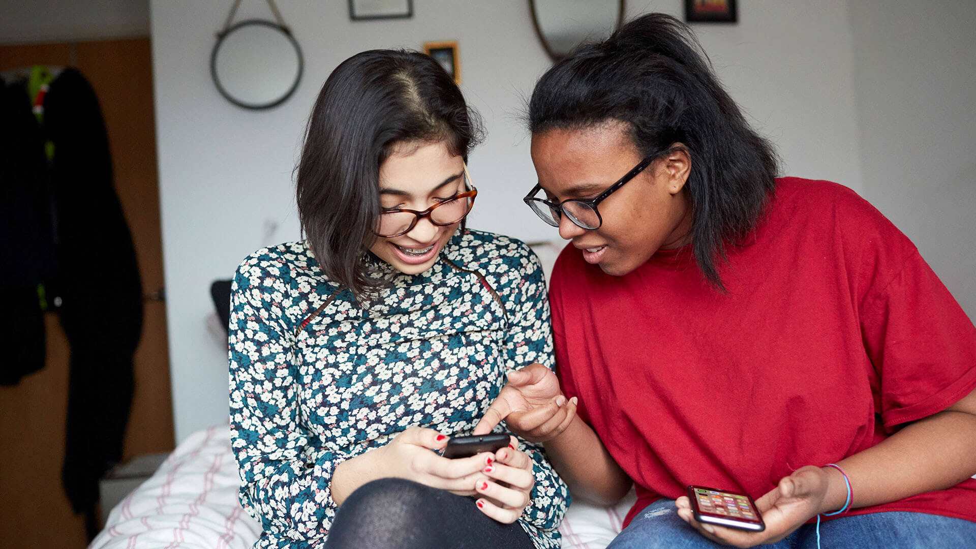 Two young people, both wearing glasses sit on a bed in a bedroom each holding a phone. The person on the right is pointing at the person on the left's phone. The person on the left is looking down at their phone and smiling.