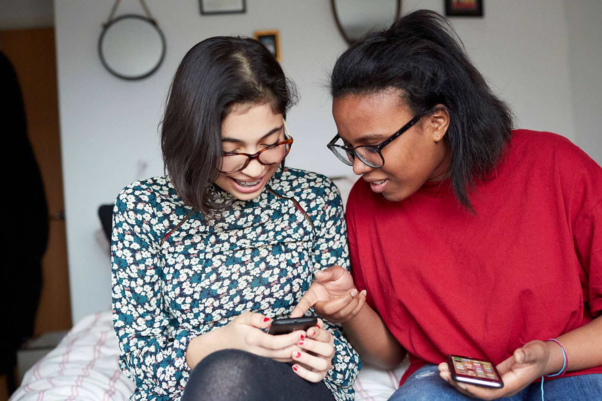 Two young people sitting together on the end of a bed and looking at their mobile phones.