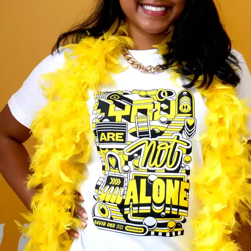 A close up of a woman smiling wearing a yellow feather bower, gold chain and our David Oku's bespoke 'You are not alone' graphic Hello Yellow T-shirt.