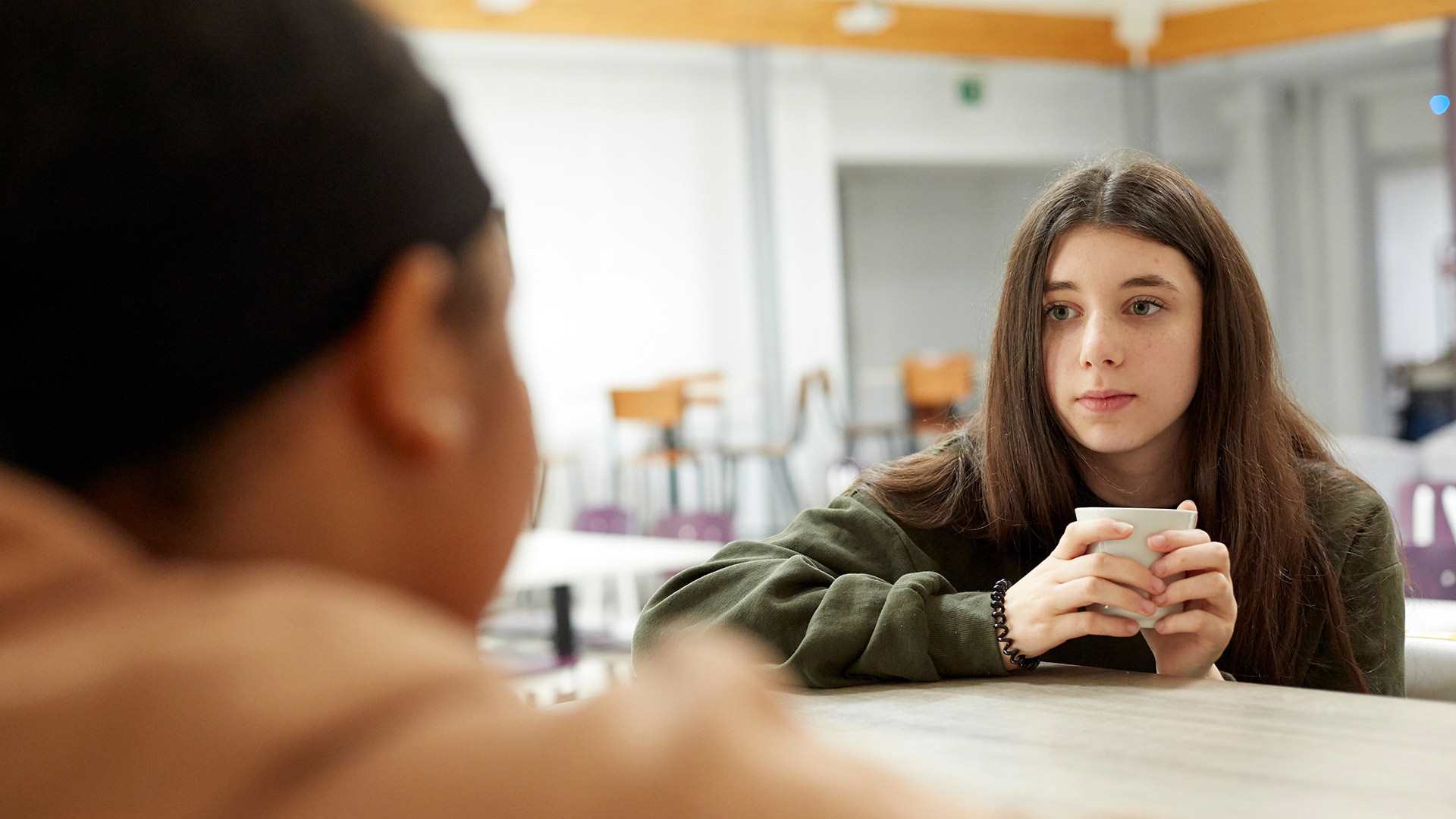 A young girl looks anxious while holding a mug and talking to her friend who is sitting opposite her in a school canteen.