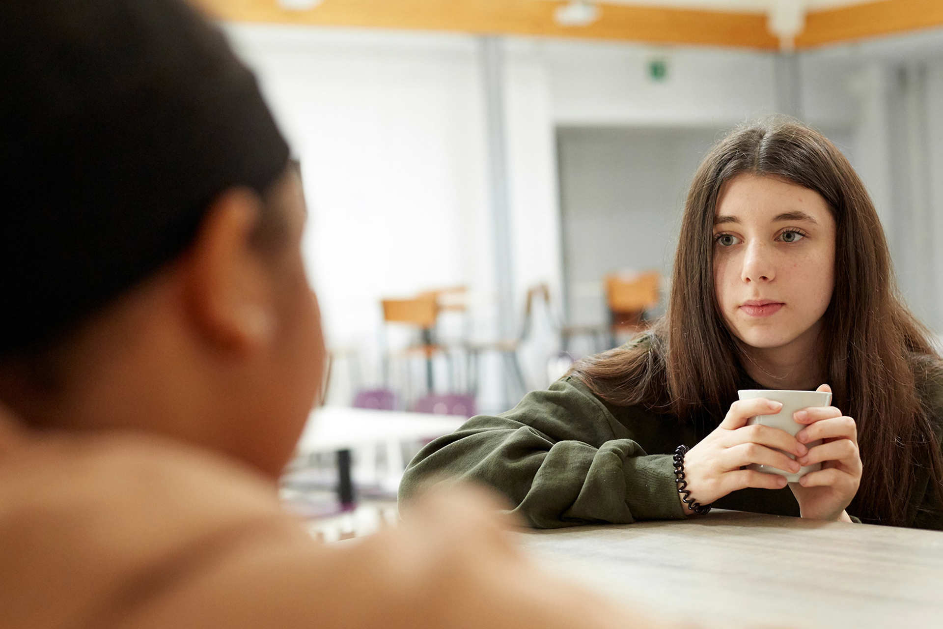 A young girl looks anxious while holding a mug and talking to her friend who is sitting opposite her in a school canteen.