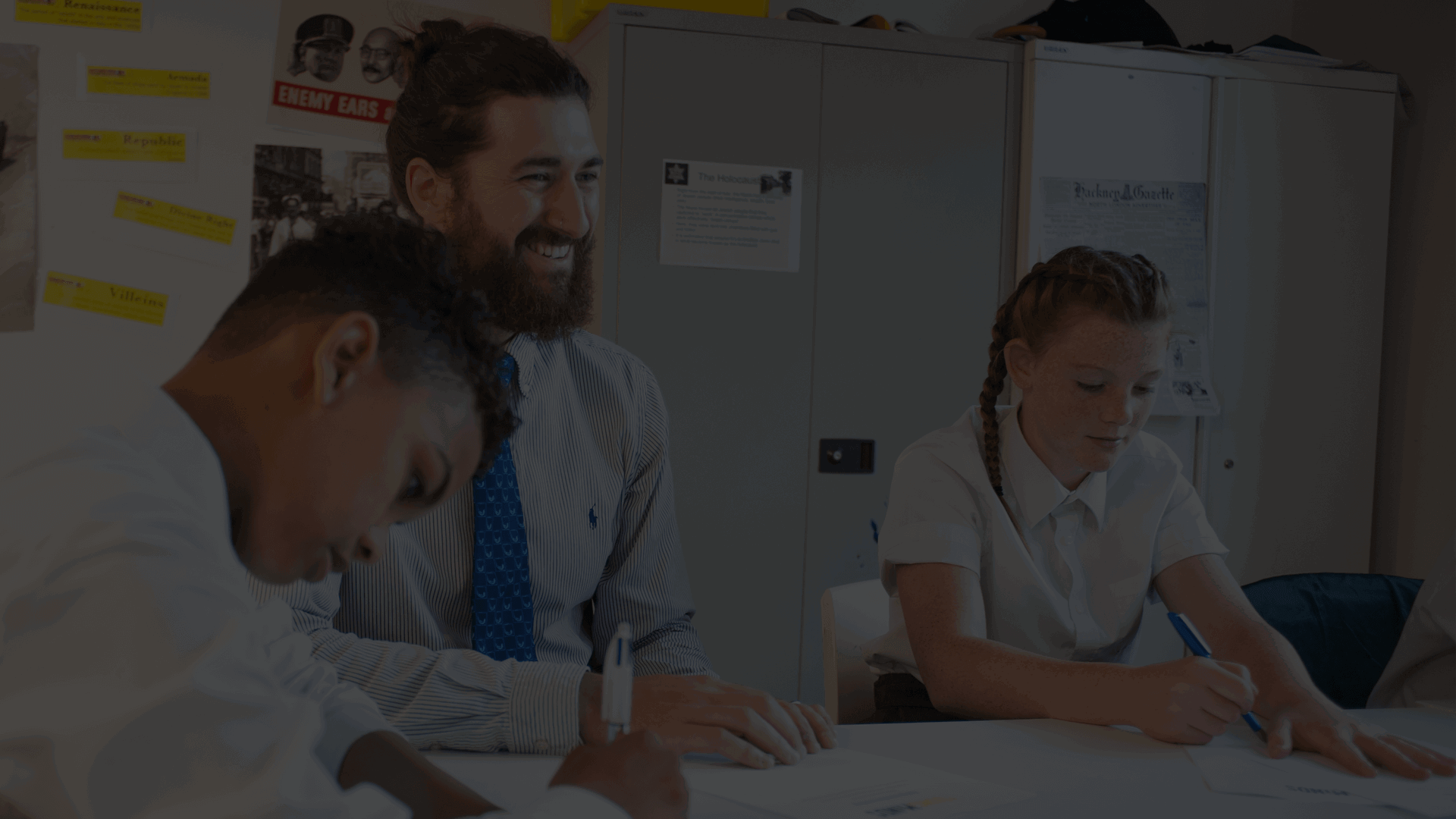 On the left side of the photo, a young person is looking down at work on a desk while holding a pen. In the middle is the teacher, looking forward and smiling. To the right of the teacher is another young person looking down and writing on paper. They are sitting in a classroom setting round a desk.